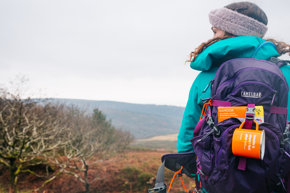 A woman on a walk wearing a waterproof coat and woolly hat. She is carrying a rucksack with a map of the area.