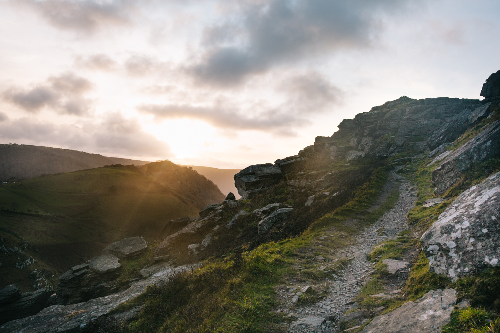 Rocky hillside path at sunset with sun rays breaking through clouds.